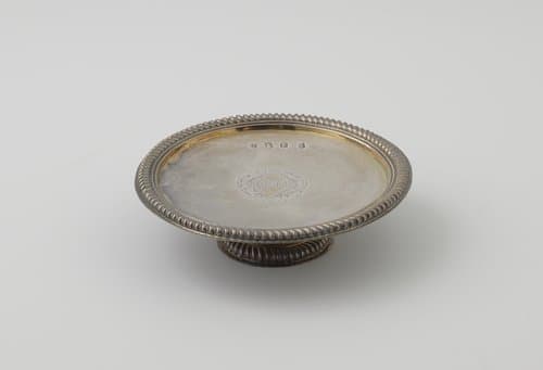 Presenteerblad van zilver, rond, langs de rand afgezet met een zware kabelrand. Op het plat een ruitvormig wapenschild tussen blad- en cartouchemotieven. Korte, ronde voet. by William Denny