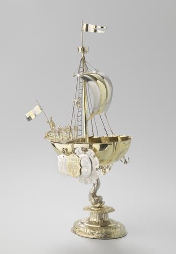 Rederijbeker van verguld zilver in de vorm van een schip gedragen door een dolfijn. Het schip is getuigd met mast, want en zeil en is bemand met 12 man.